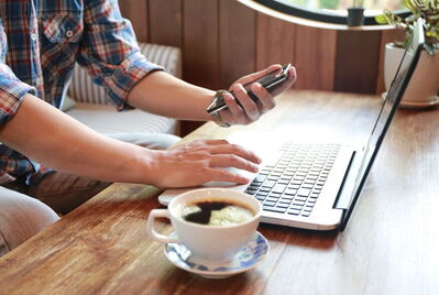 Person sat at the coffee table typing on their laptop whilst also on their phone