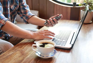 Person typing into a laptop and looking at a mobile phone